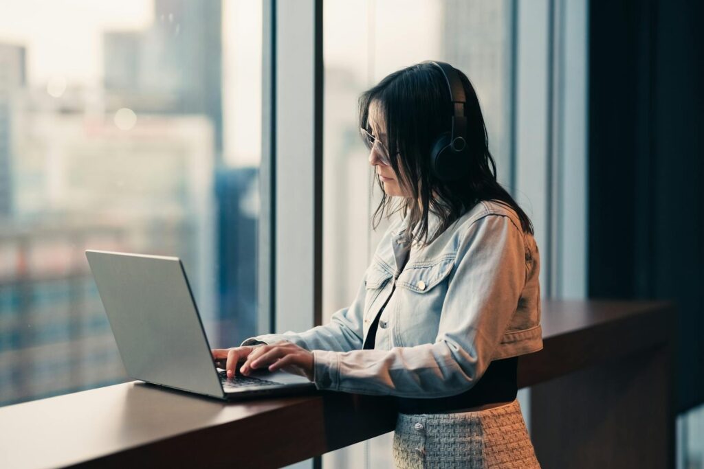 A woman wearing headphones works on her laptop in a high-rise building, looking out a large window with a city view.